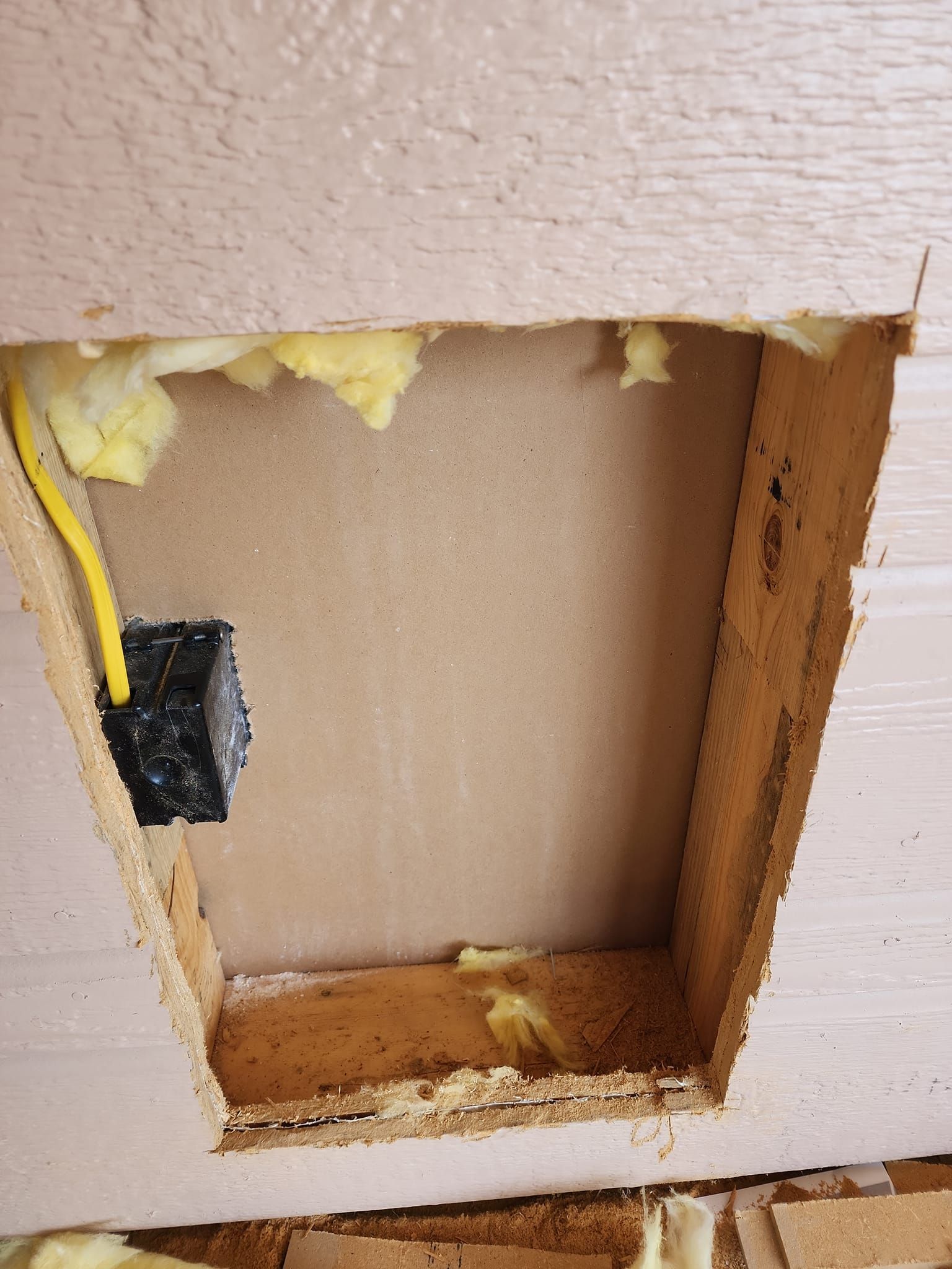 Open wall cavity exposing electrical box and insulation. Beige siding surrounds the opening.