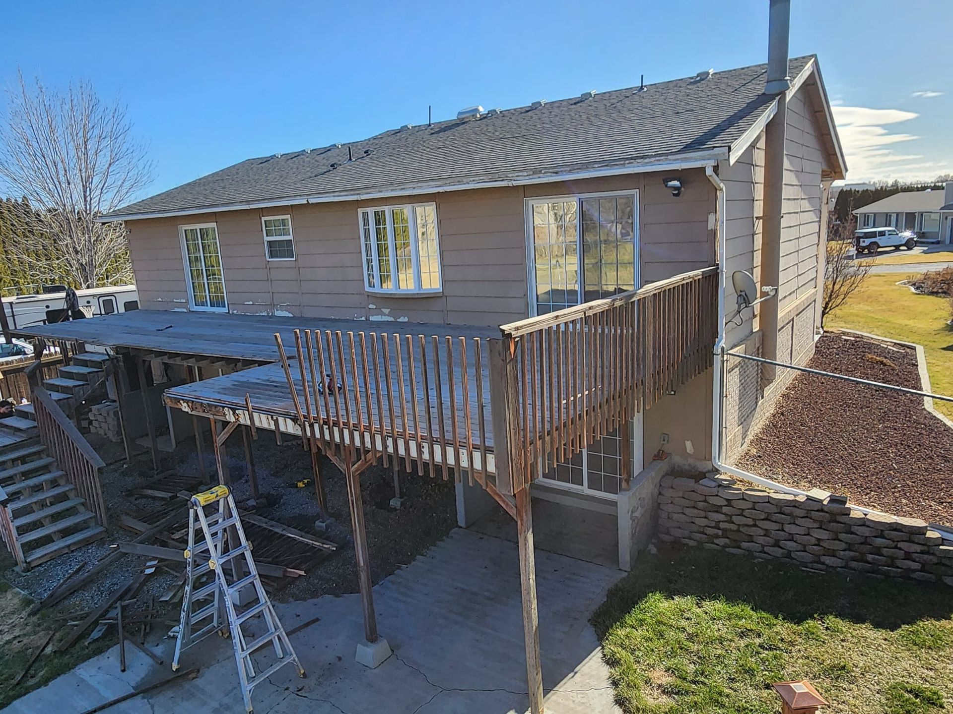 Two-story house with a wooden deck and a lower-level patio. The house is tan, with a chimney. A ladder is present.