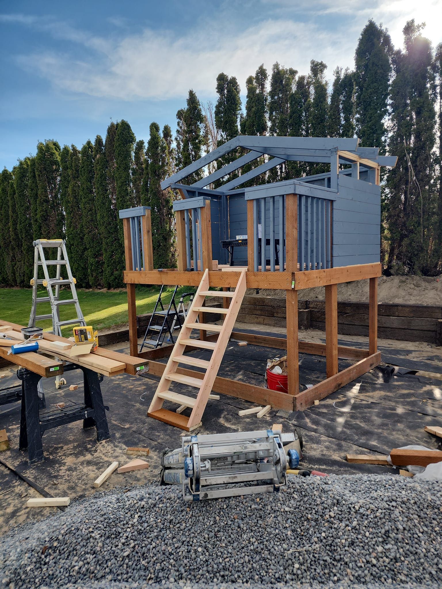 Wooden playhouse under construction with steps, gray paint, and surrounding landscaping.