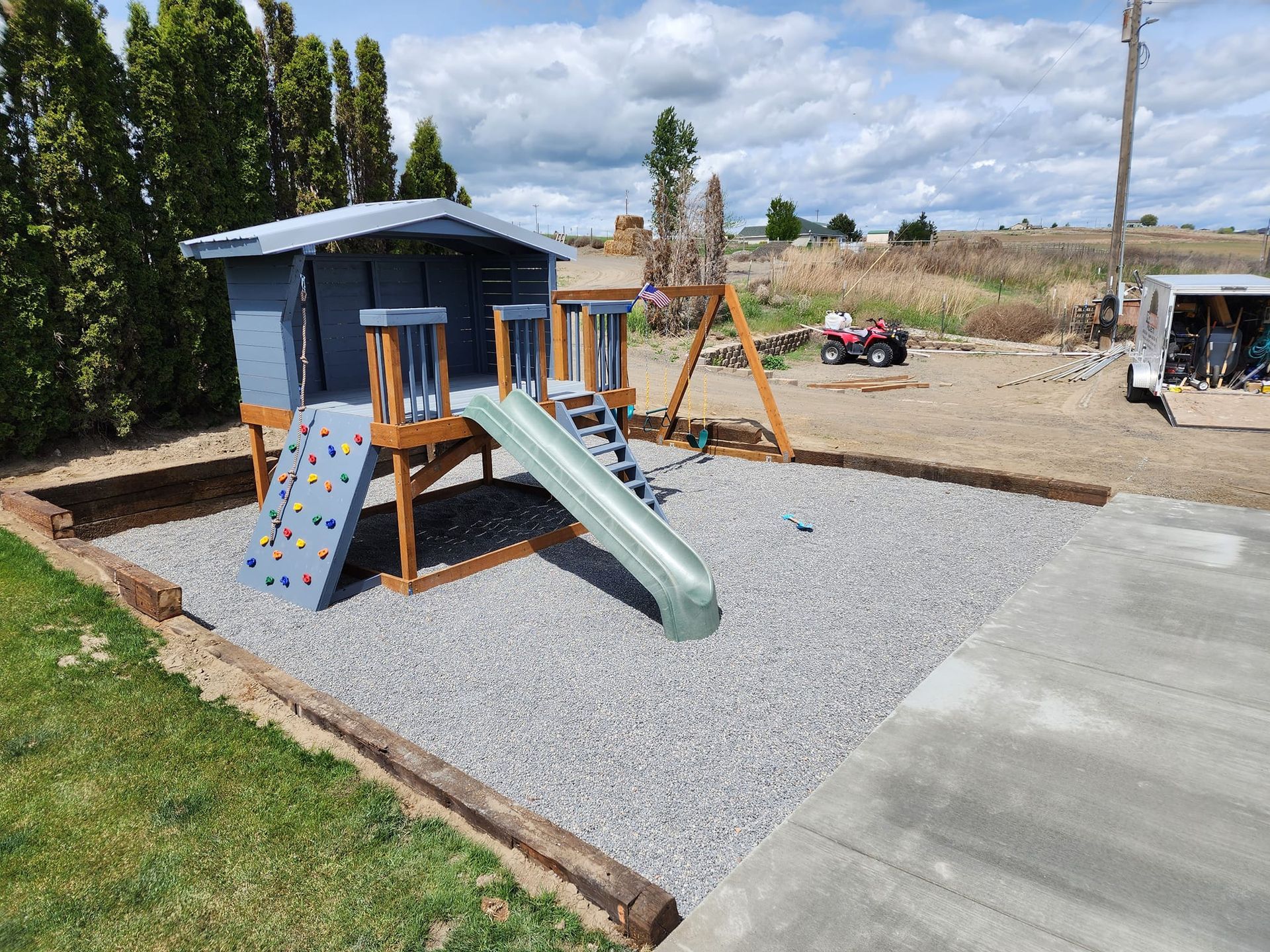 Playground with gray house, slide, climbing wall, swings, and gravel ground.