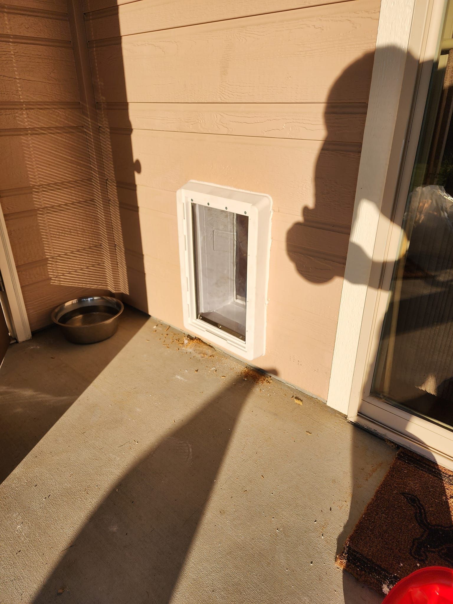 Pet door in a wall with a dog bowl nearby. Brown siding, white trim, and a concrete patio.