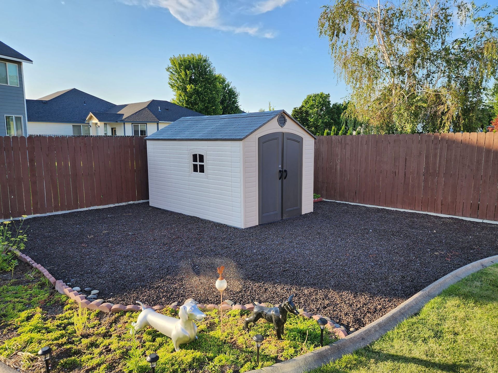 A small storage shed in a gravel area within a fenced backyard, with statues in the foreground.