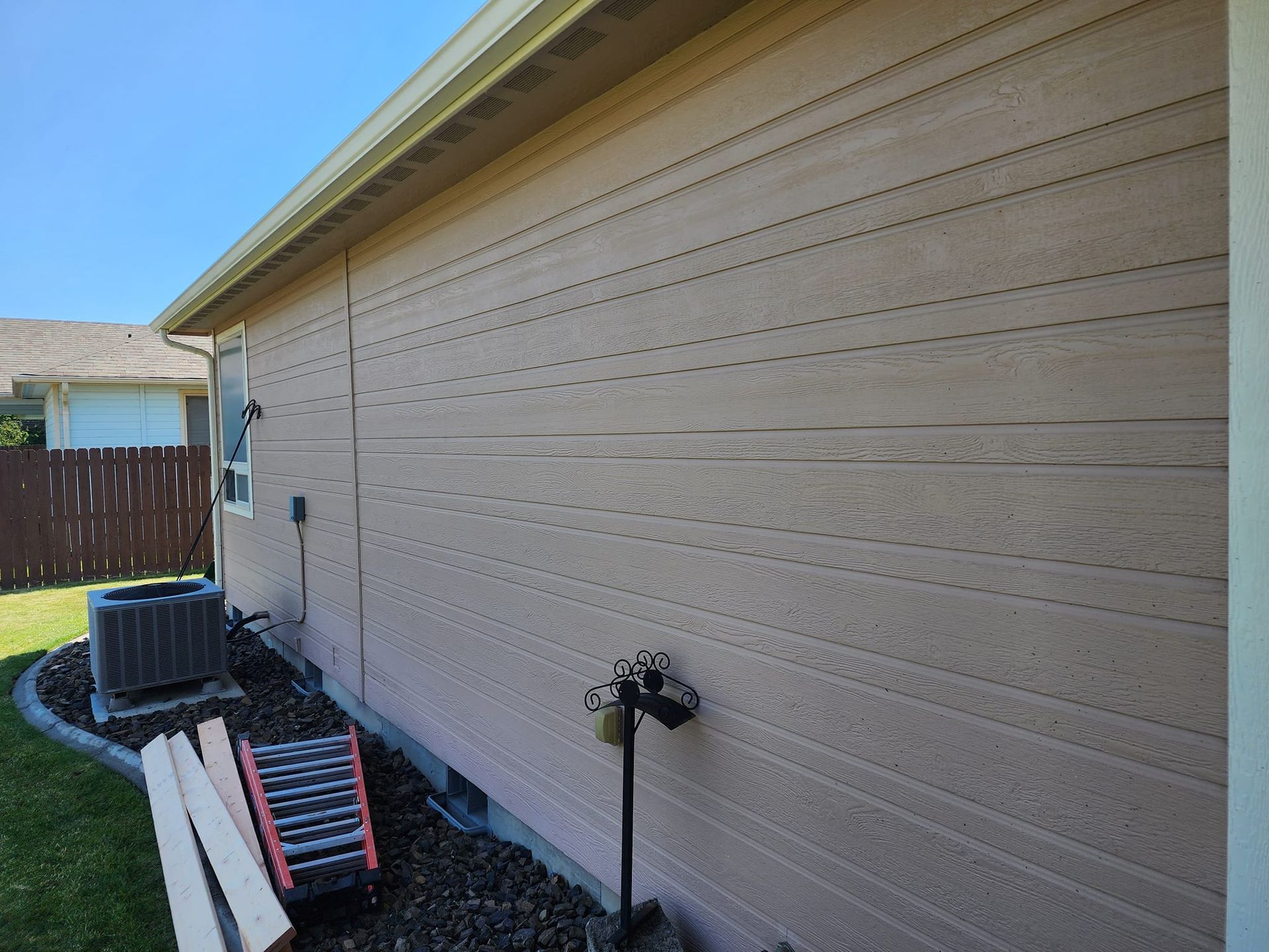 Exterior wall of a house with light brown siding, gutter, and a fence in the background.