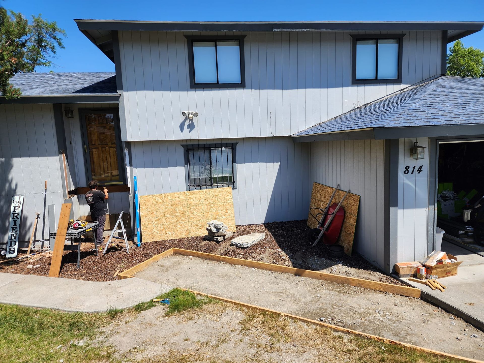 House exterior with ongoing construction. Person working on front porch. Brown siding, blue roof, sunny day.