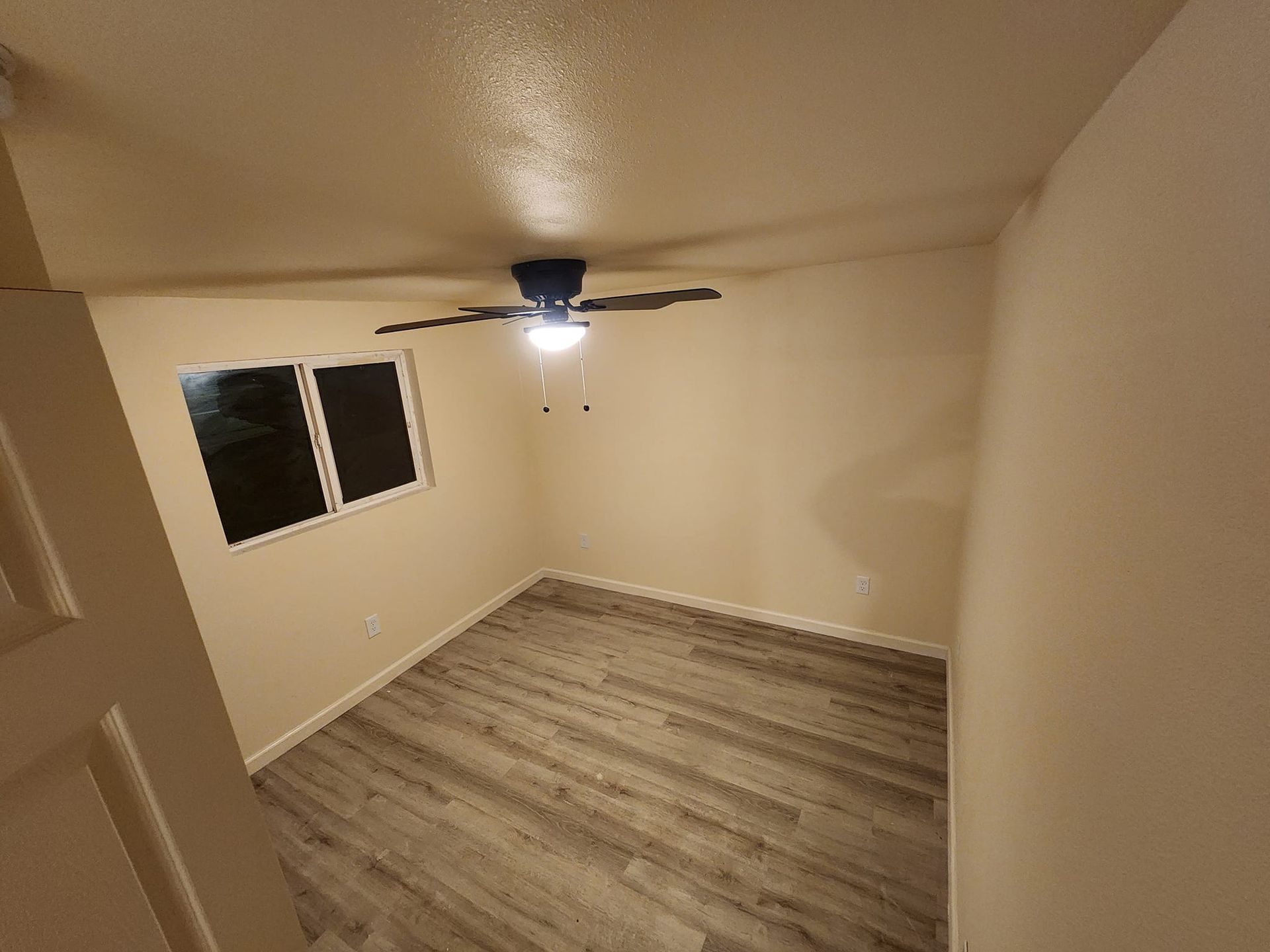 Empty room with gray wood-look flooring, light walls, and a ceiling fan. Window on the left, door on the left.