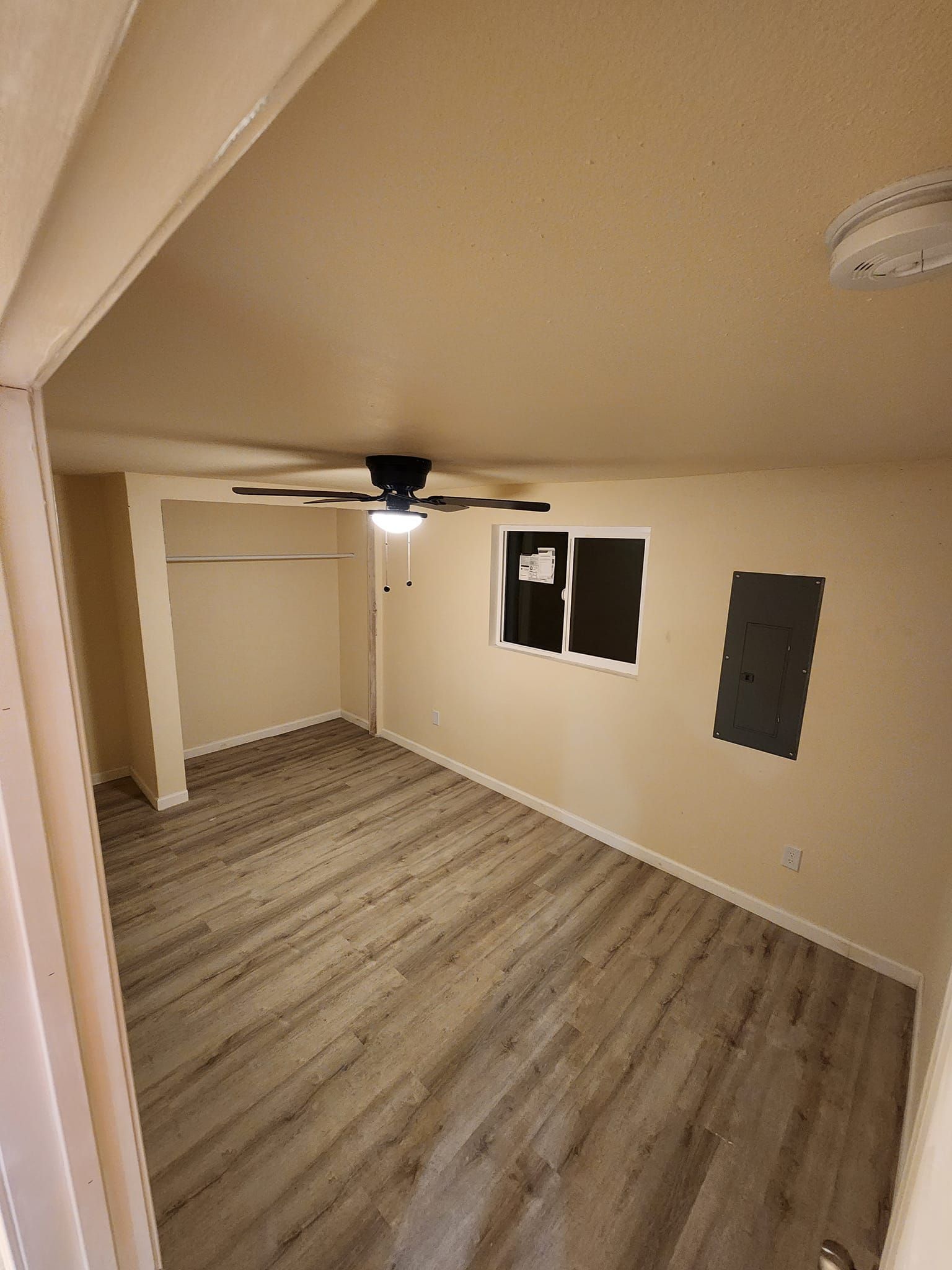 Empty bedroom with wood-look flooring, beige walls, closet, ceiling fan, and window.