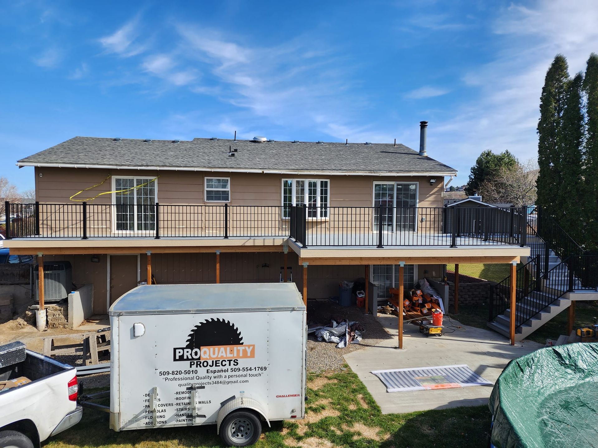 House with two-level deck being constructed, blue sky overhead, trailer and equipment in yard.
