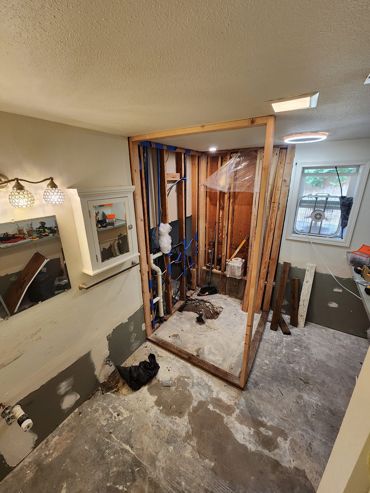 Bathroom under renovation; exposed framing outlines shower, pipes visible. Gray walls, debris, and bright ceiling lights.
