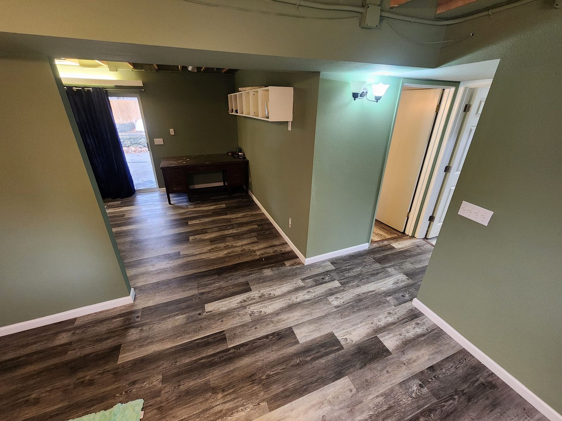 Dark wood-look flooring in a room painted olive green, leading to a doorway and hallway, with light fixtures.