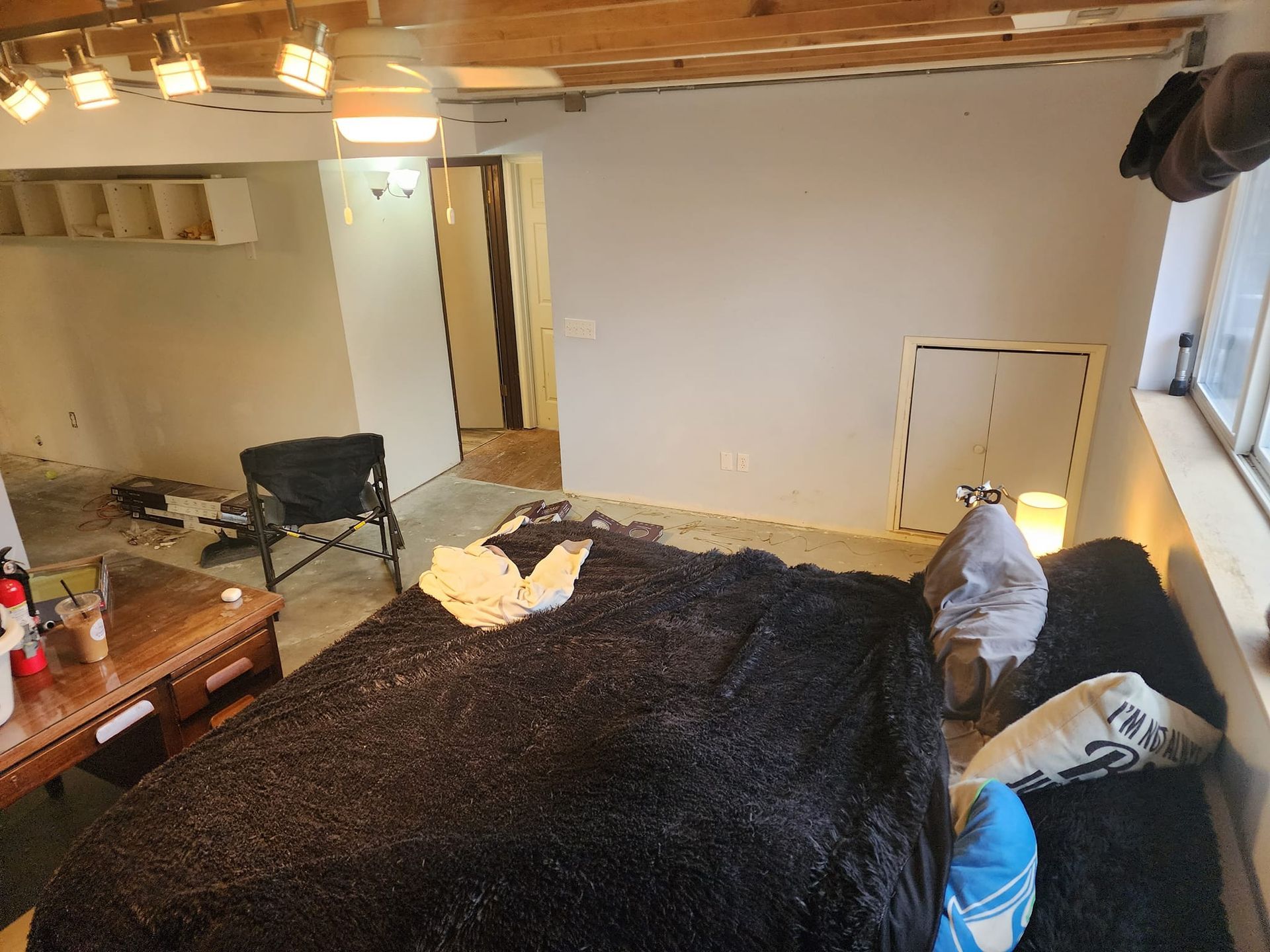 A cluttered basement bedroom with a black fuzzy bed, a small table, and a window.