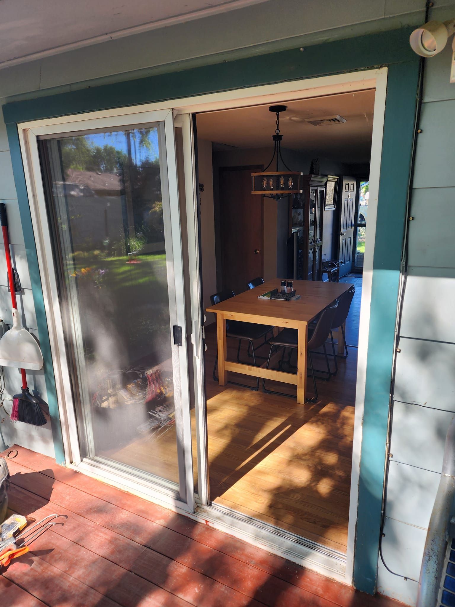 Sliding glass door open, leading from a deck to a kitchen with a wooden table. Sunlight streams in.