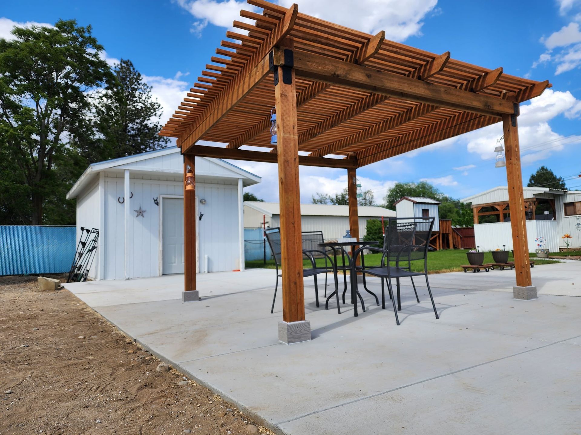 Wooden pergola over concrete patio with table and chairs, white building in background, blue sky.