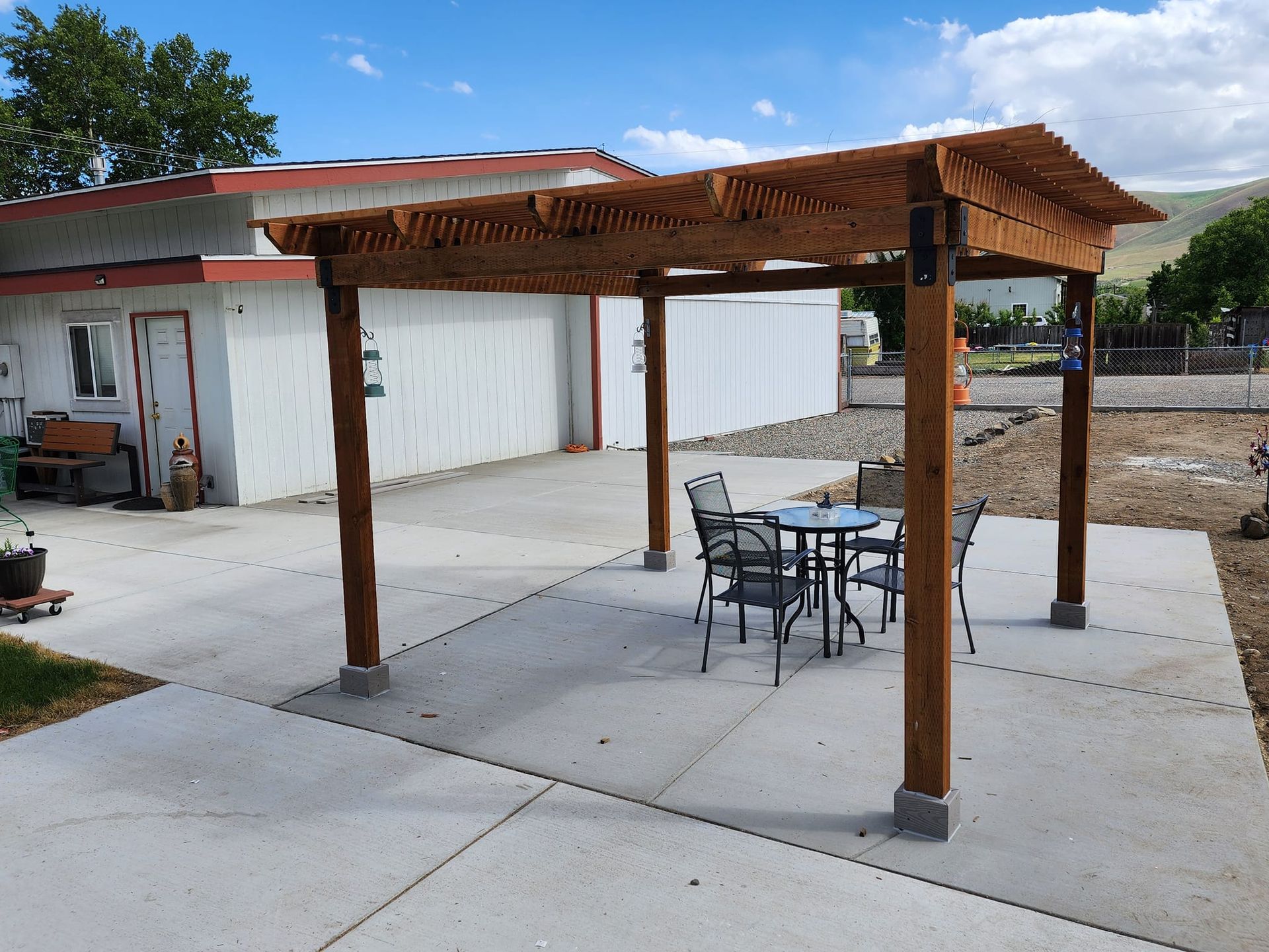 Wooden pergola with a table and chairs on a concrete patio in front of a white building.