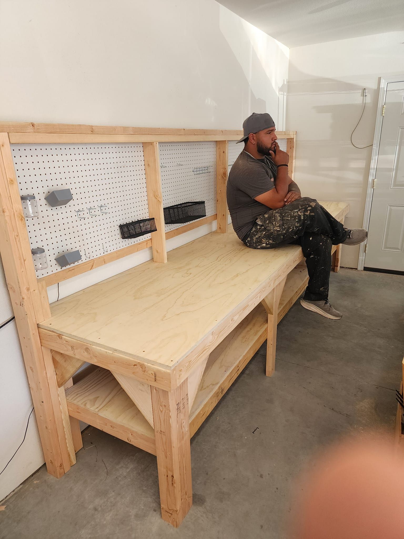 Man sitting on a wooden workbench in a garage, looking thoughtful.