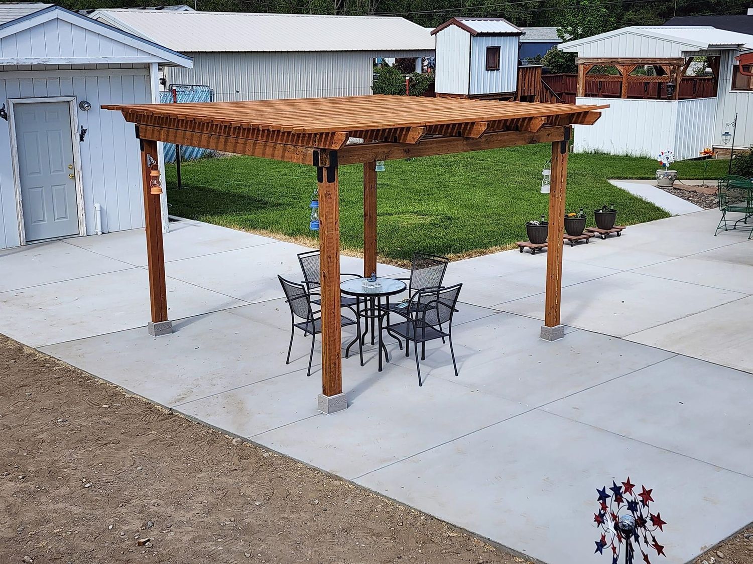 Wooden pergola over a patio with a table and chairs, set against a green lawn and white buildings.