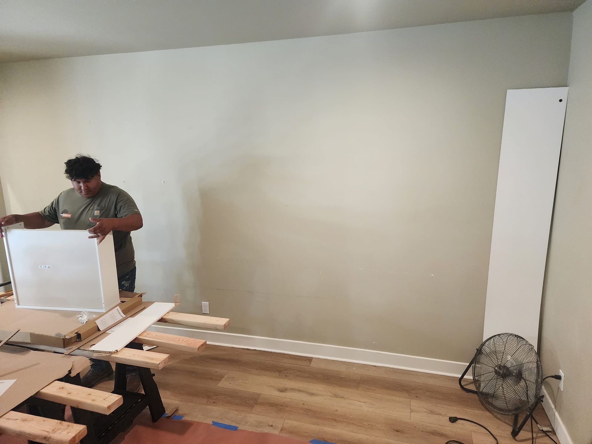 Person measuring and assembling white cabinet components against a light-colored wall, with a fan nearby.