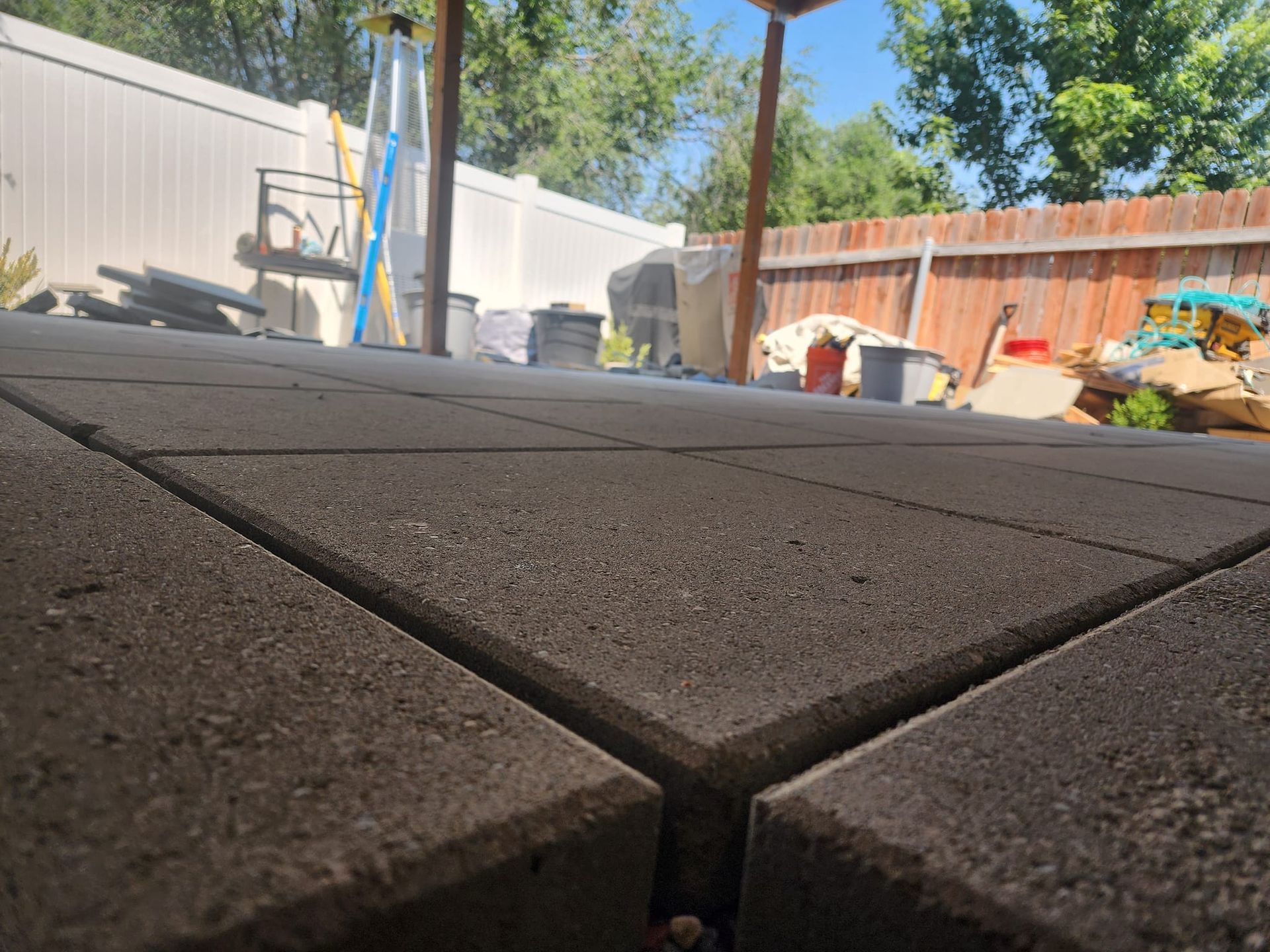 Low-angle view of gray paver patio with visible lines. A wooden structure, fence, and various items are in the background.