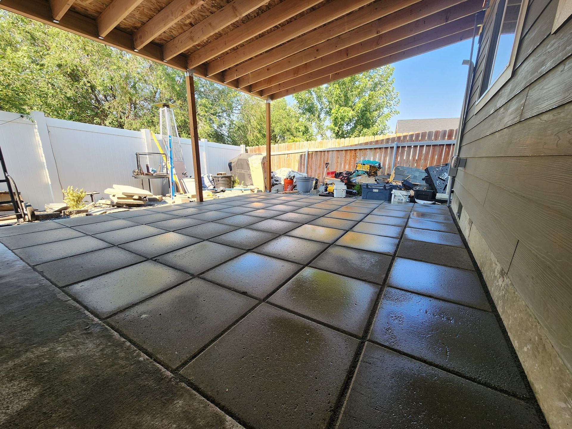 Paved patio beneath a covered wooden deck, bordered by a white fence and a house with green siding; various items present.