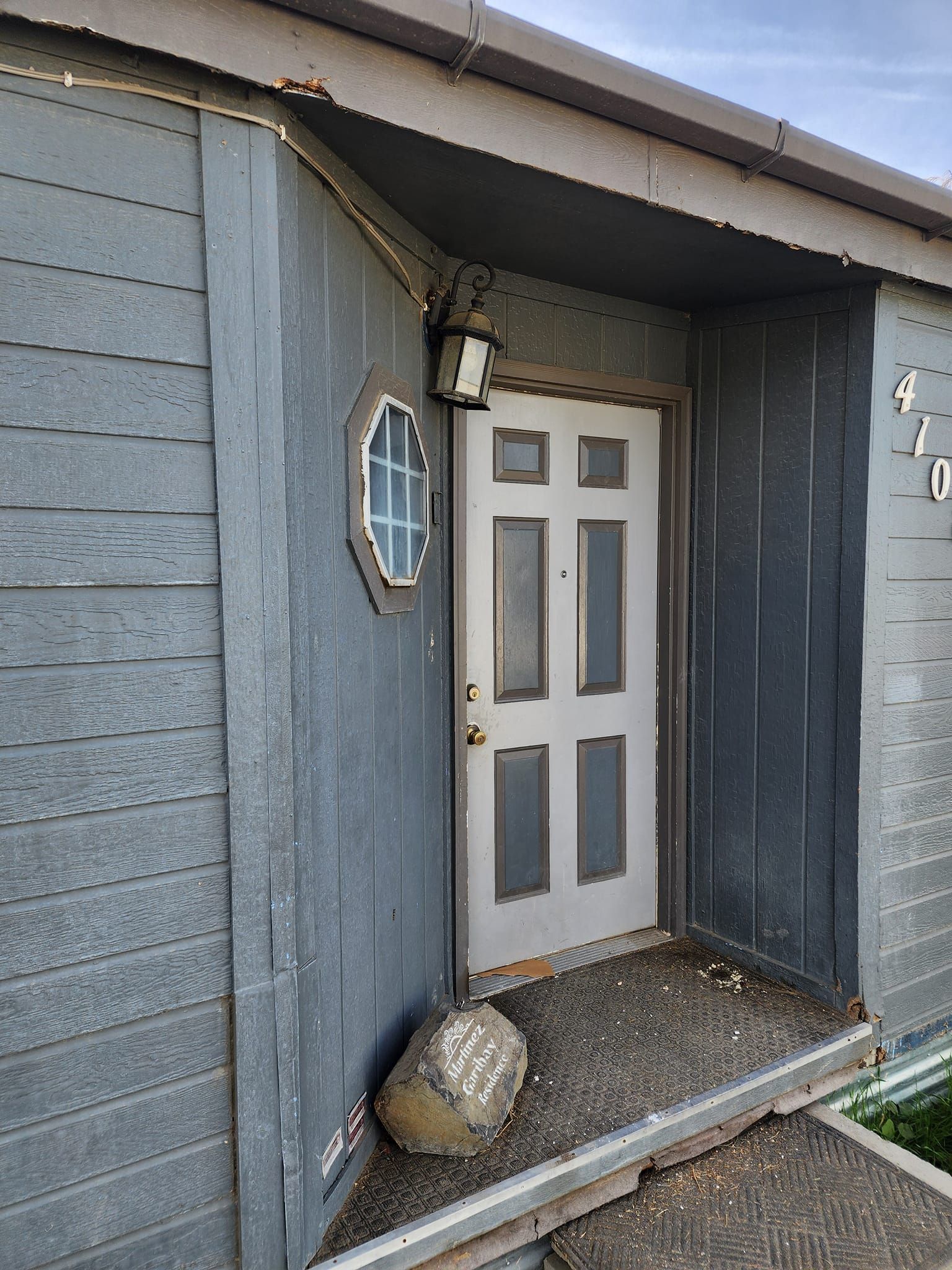 Gray wooden house front entrance with a light fixture, round window, and rock on the porch.