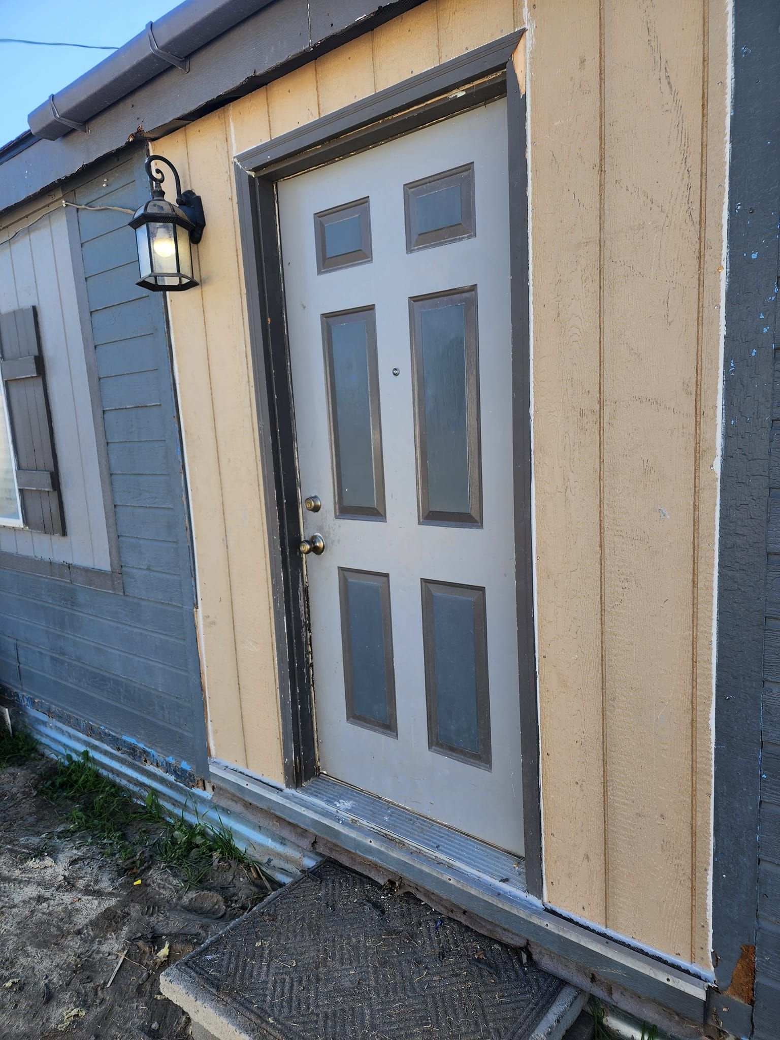 Exterior view of a small building with a light gray door, tan trim, and a black exterior light.