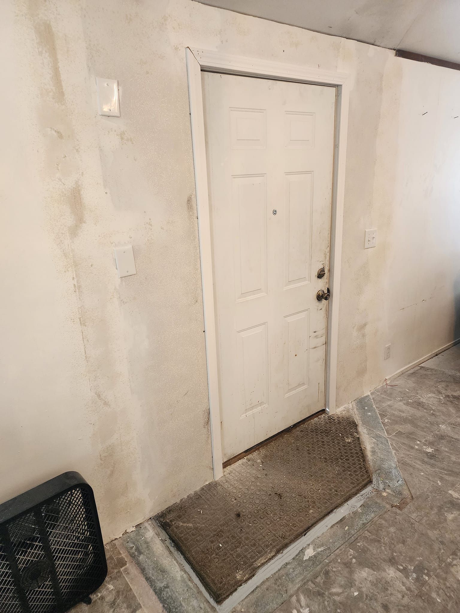 White door in doorway; water-stained wall. Black fan on the floor. Concrete and rug.