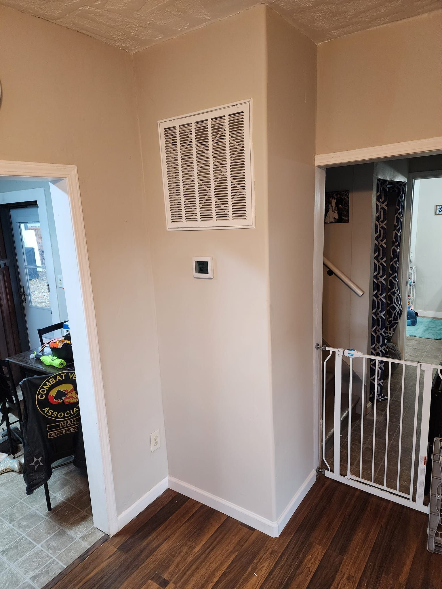 Corner of a room with air vent, thermostat, and doorways. Wooden floor, beige walls, and a white safety gate.