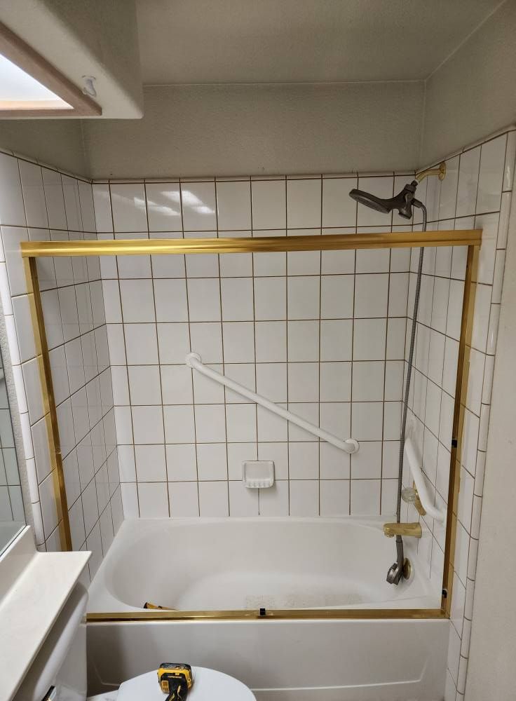 Bathtub with white tile walls and a gold-framed glass shower door. A grab bar is mounted on the wall.