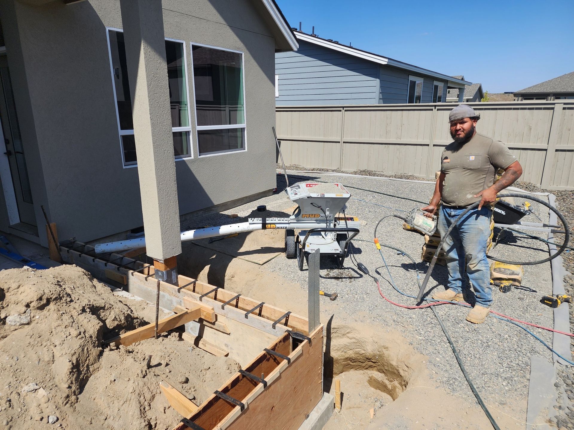 Construction worker stands near a house, next to a saw, concrete forms, and a trench on a sunny day.