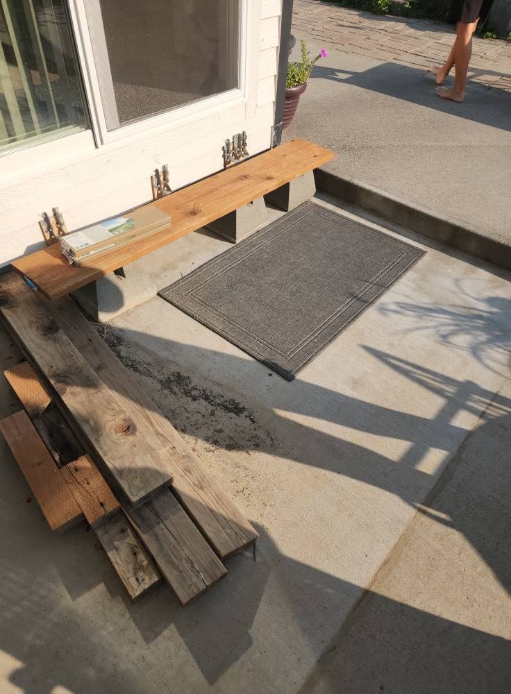 Wooden bench, rug, and stacked wood planks by a doorway. Concrete patio, sunny outdoor setting.