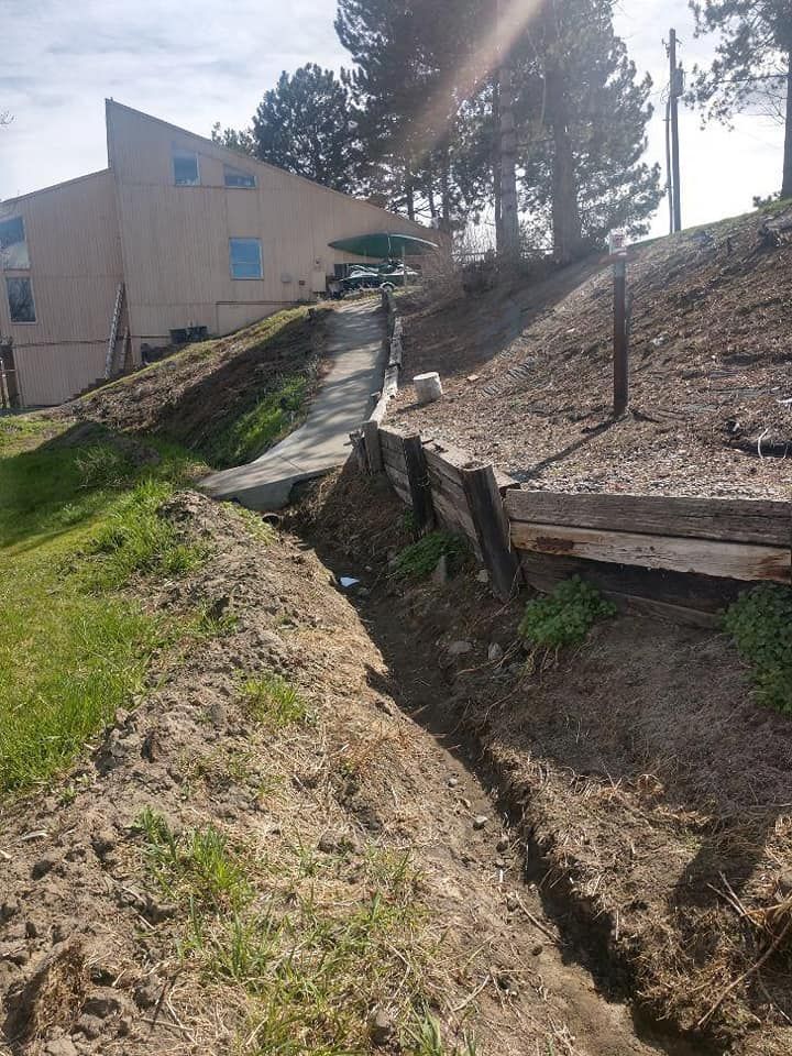 Dug trench along a dirt path leading uphill towards a building, with wooden supports on the sides.