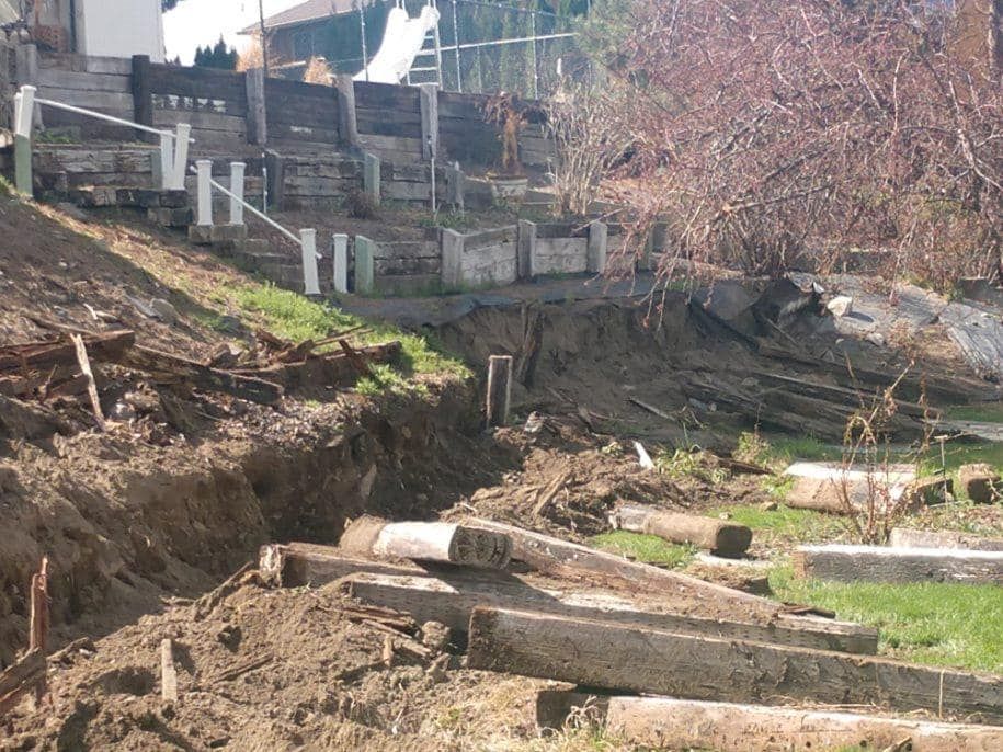 A hillside with damaged concrete steps and retaining walls, showing erosion and debris.