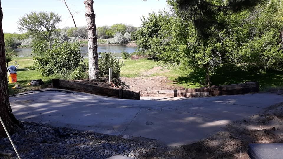 Concrete boat ramp leading to a river, surrounded by trees and greenery under a sunny sky.