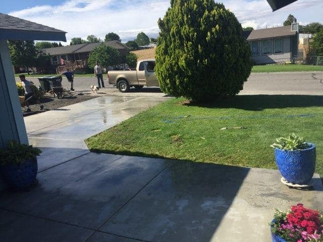People working on a driveway, truck parked nearby, green lawn, blue planters, and a tree in a residential area.