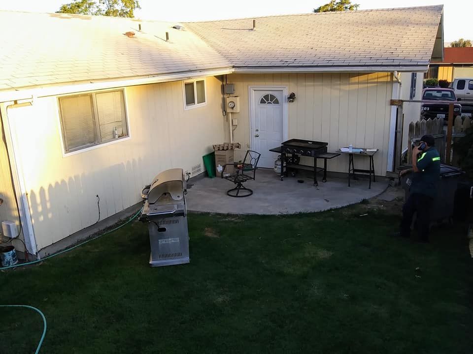 Backyard with a yellow house and patio. A person works near the fence. A grill sits on the grass.