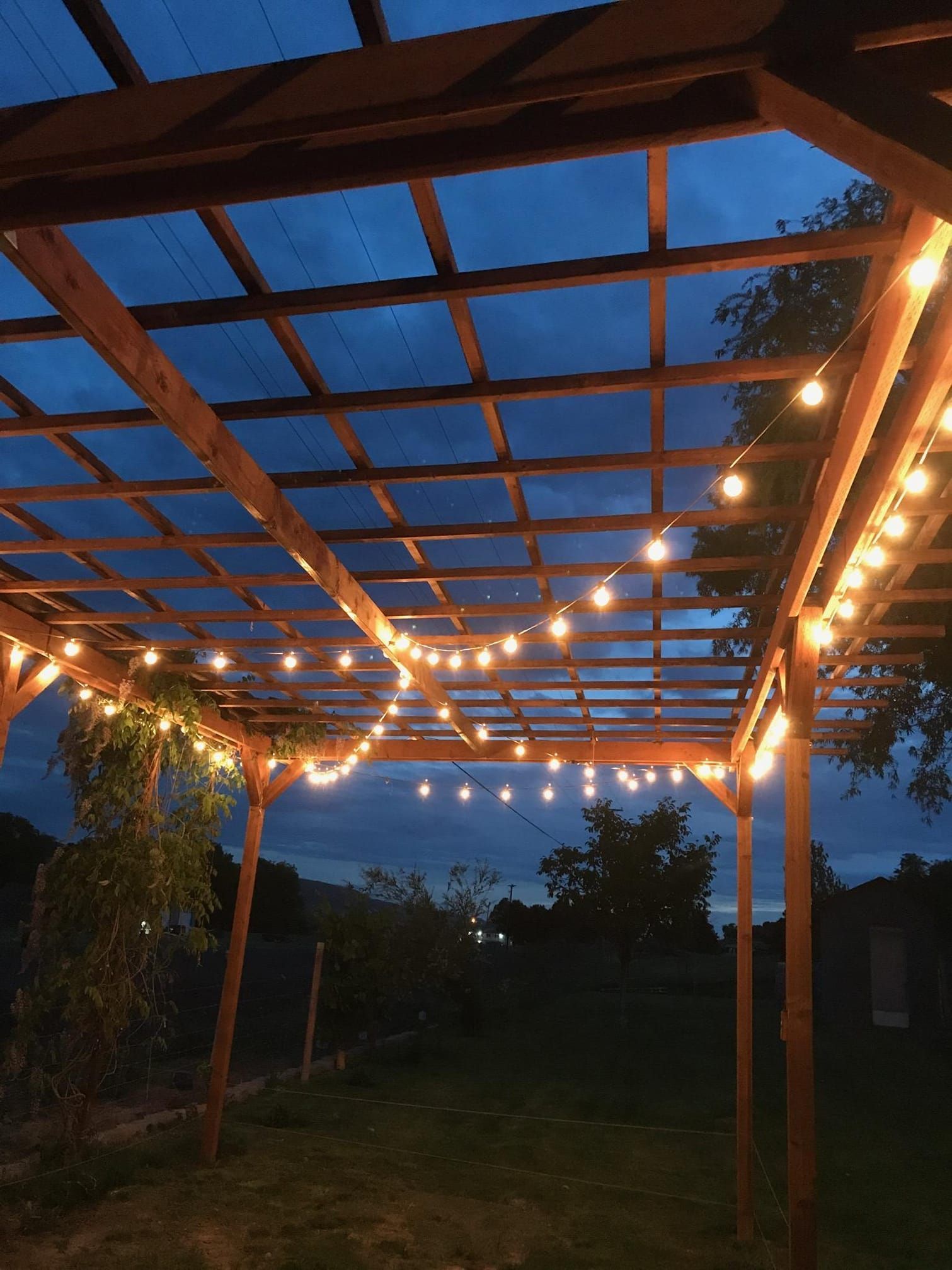 Wooden pergola with string lights, against a dusk sky, in a yard.