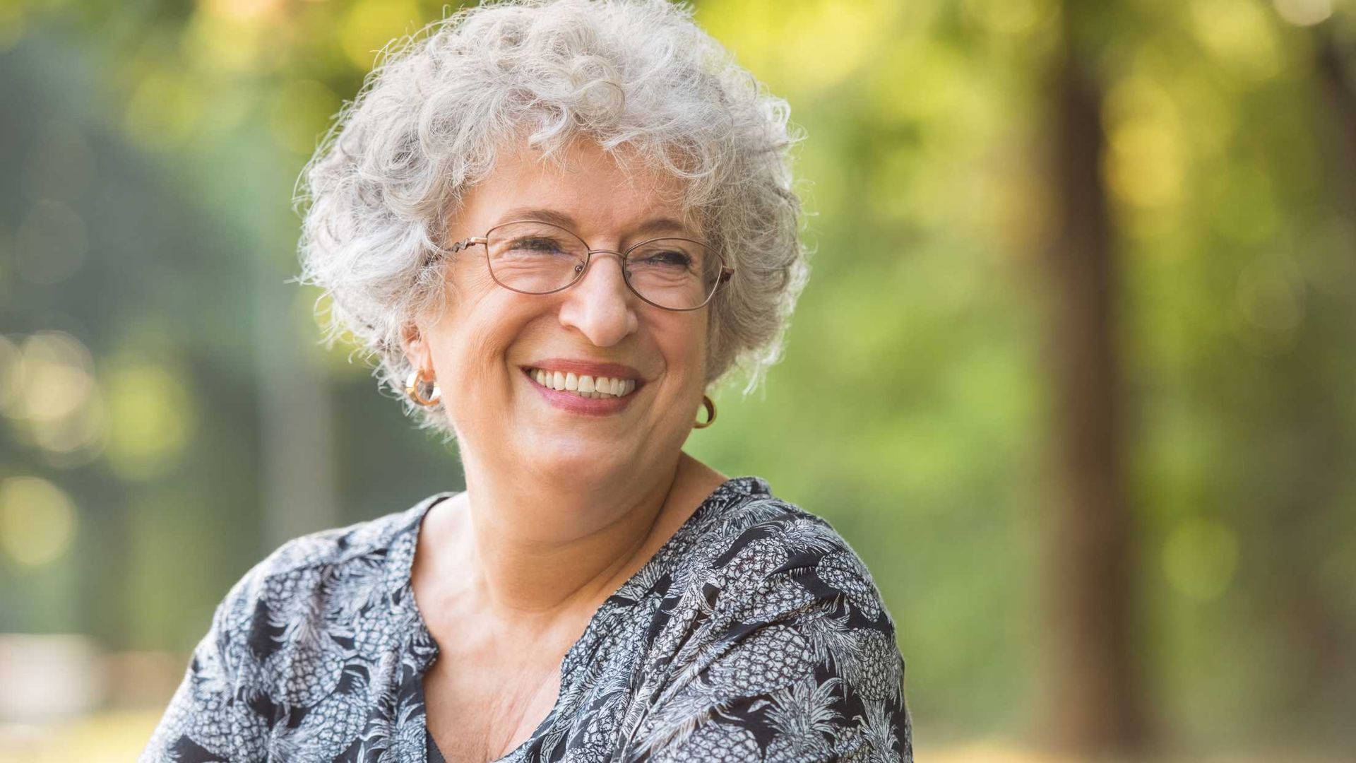 An older woman with glasses is smiling in a park.