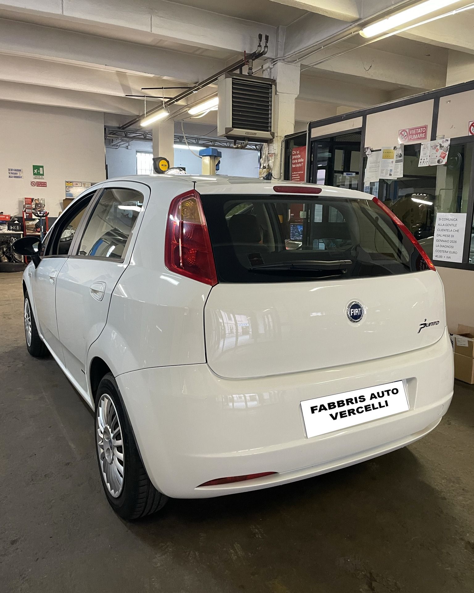 Una compatta bianca a due volumi parcheggiata all'interno di un'officina di una concessionaria, con i documenti di revisione esposti sul lunotto posteriore.
