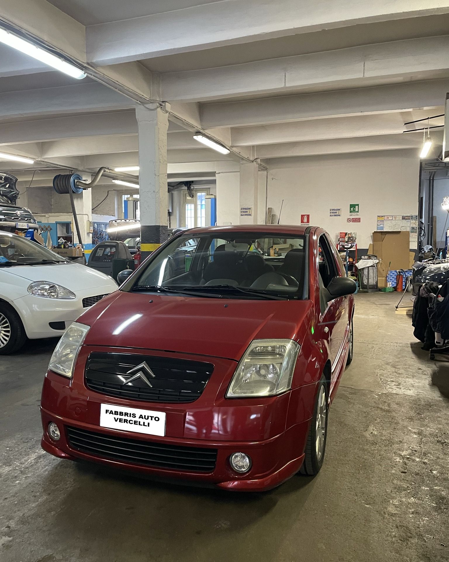 Un'auto Citroën rossa parcheggiata all'interno di un'officina, insieme ad altri veicoli e con le luci accese.