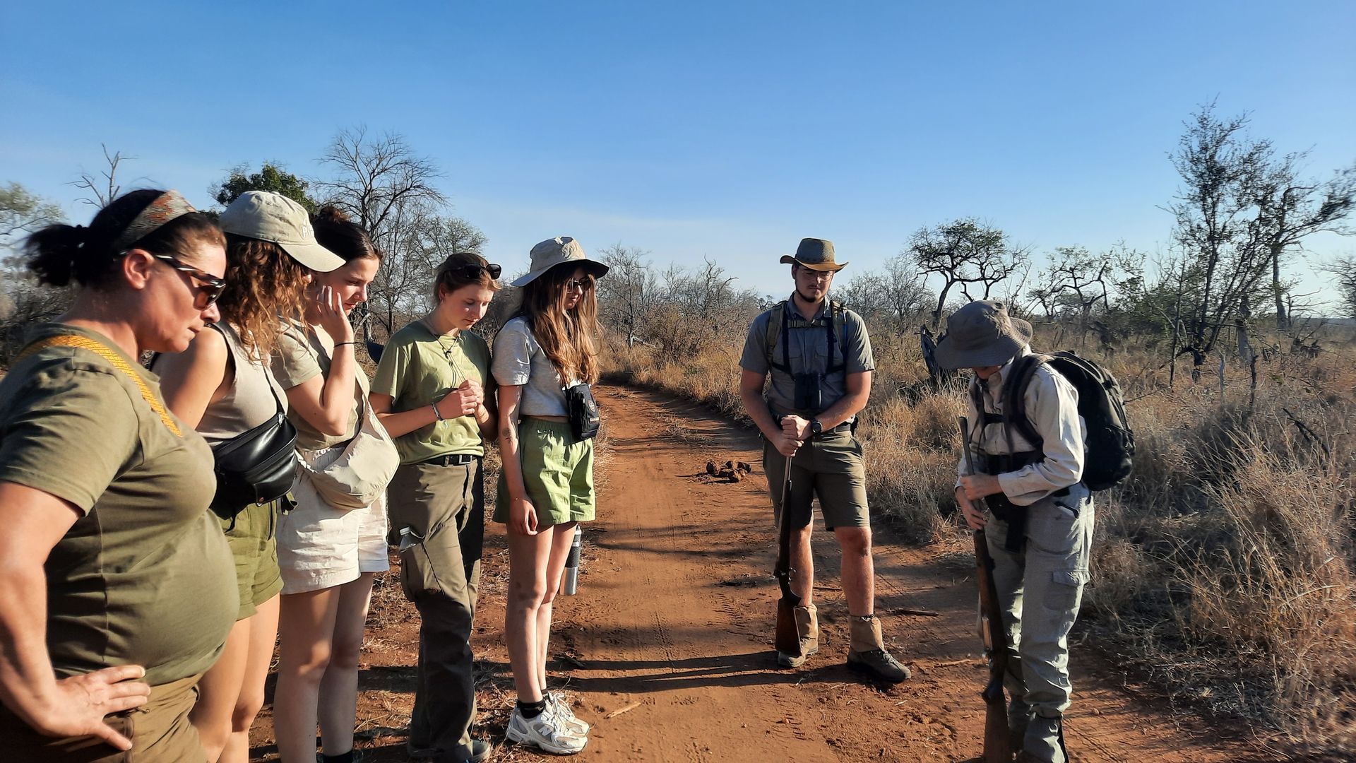 Group of people on a dirt path in a sunny savanna, listening to a guide who is pointing.