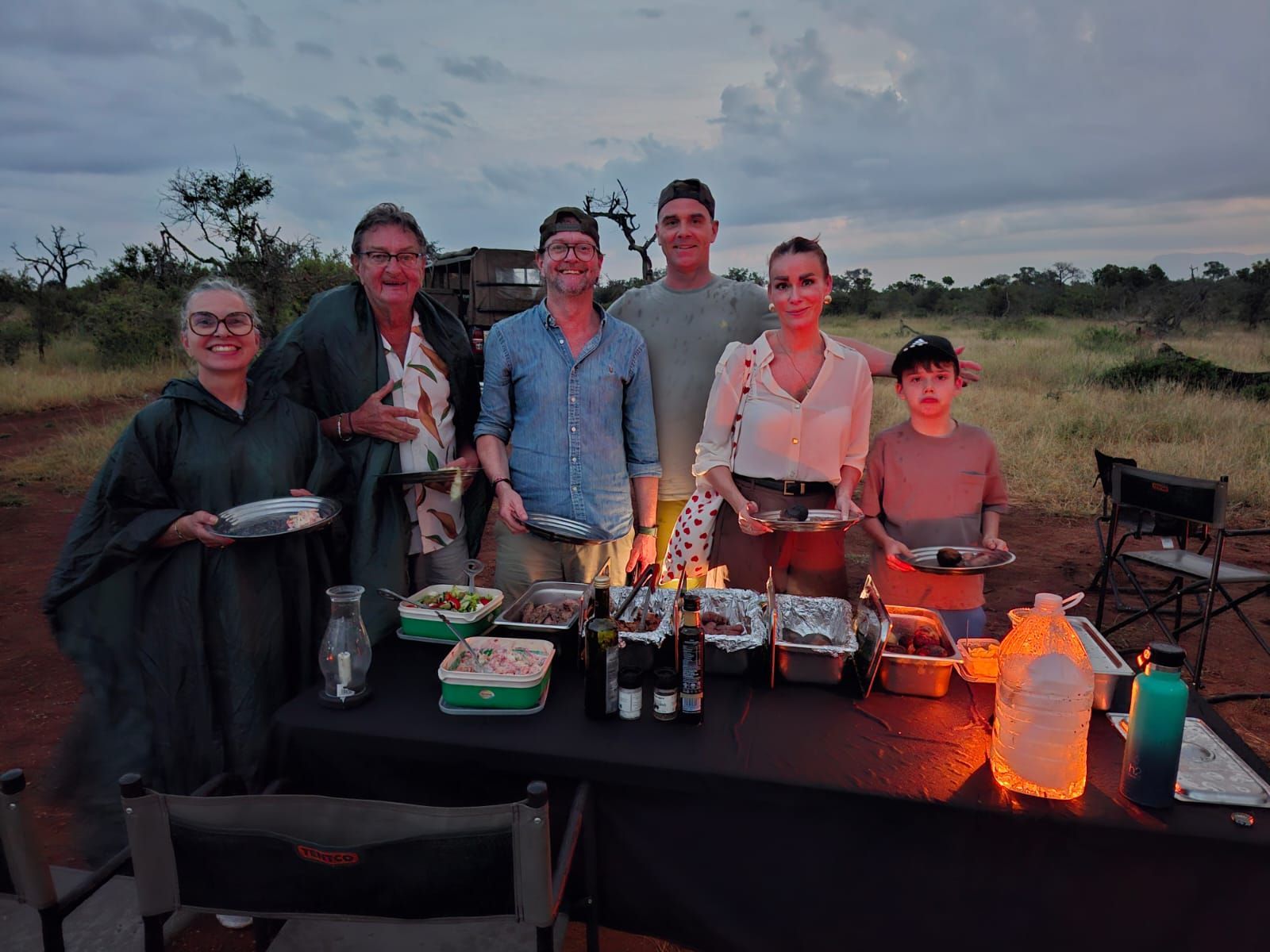 Group gathered behind a table with drinks and food at dusk in an open field, with glowing lanterns and chairs.