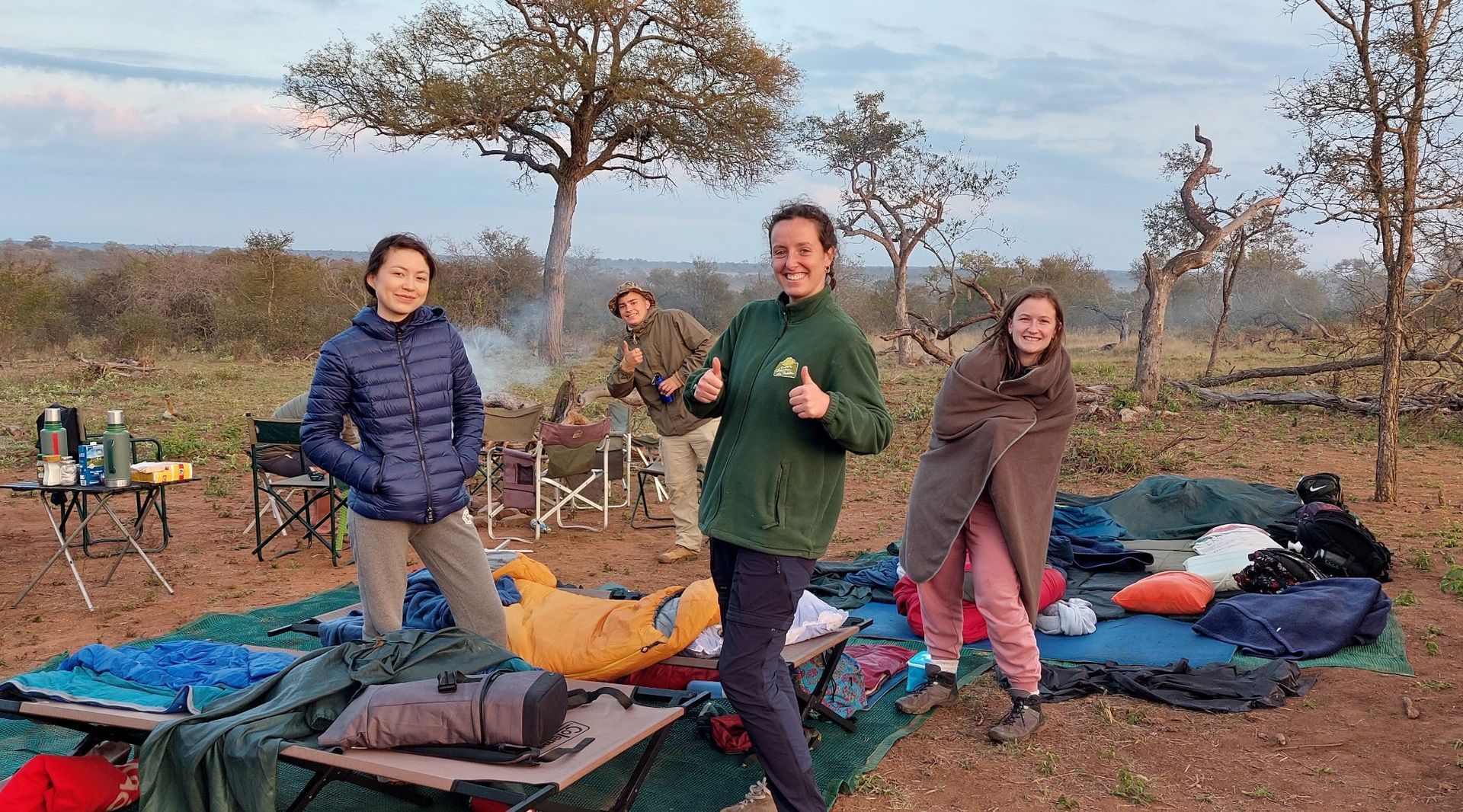 Four people stand near a campfire and camping gear in a sparse, wooded landscape during the early morning.