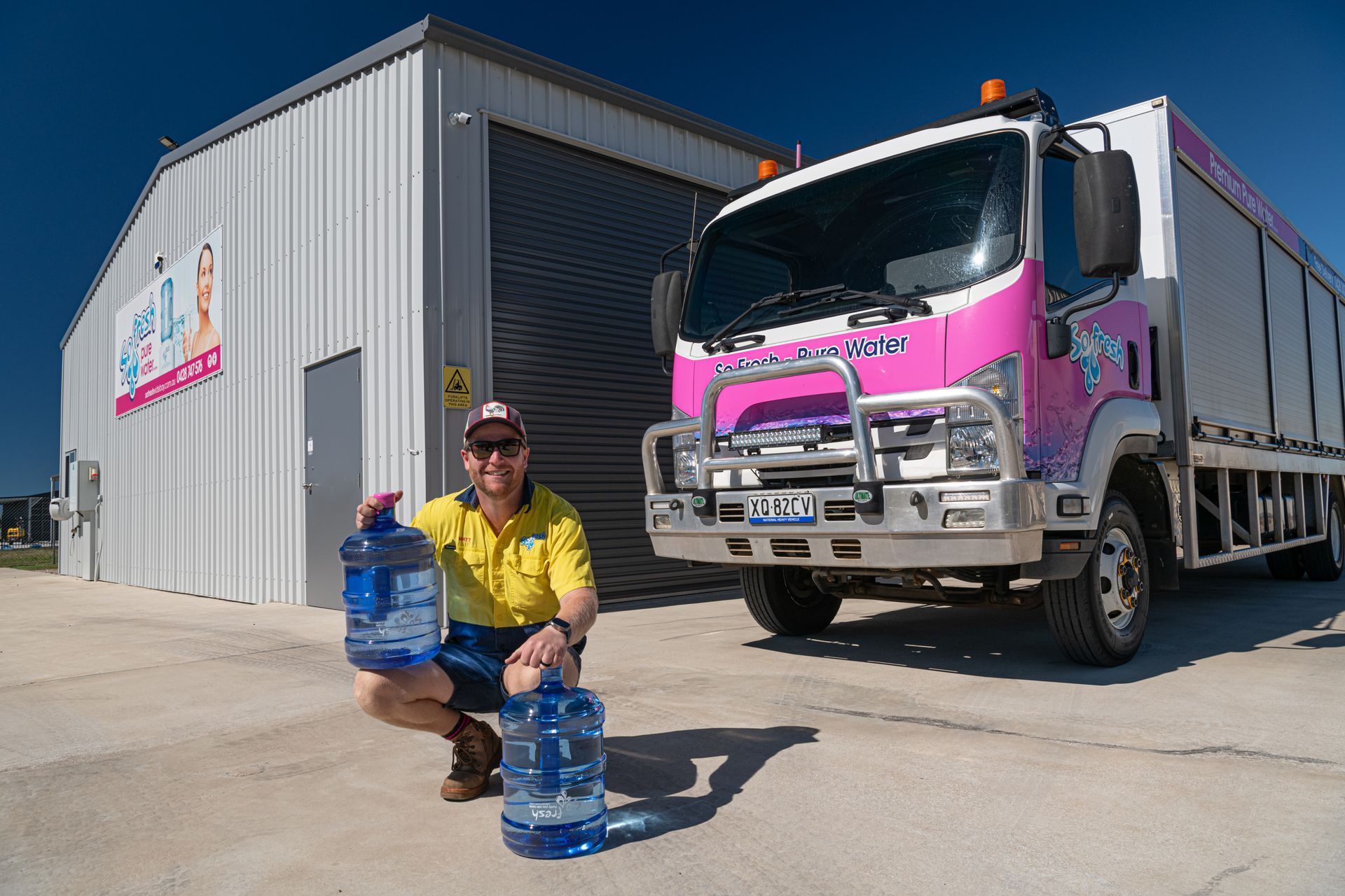 Worker crouches by water jugs beside a pink truck outside a warehouse