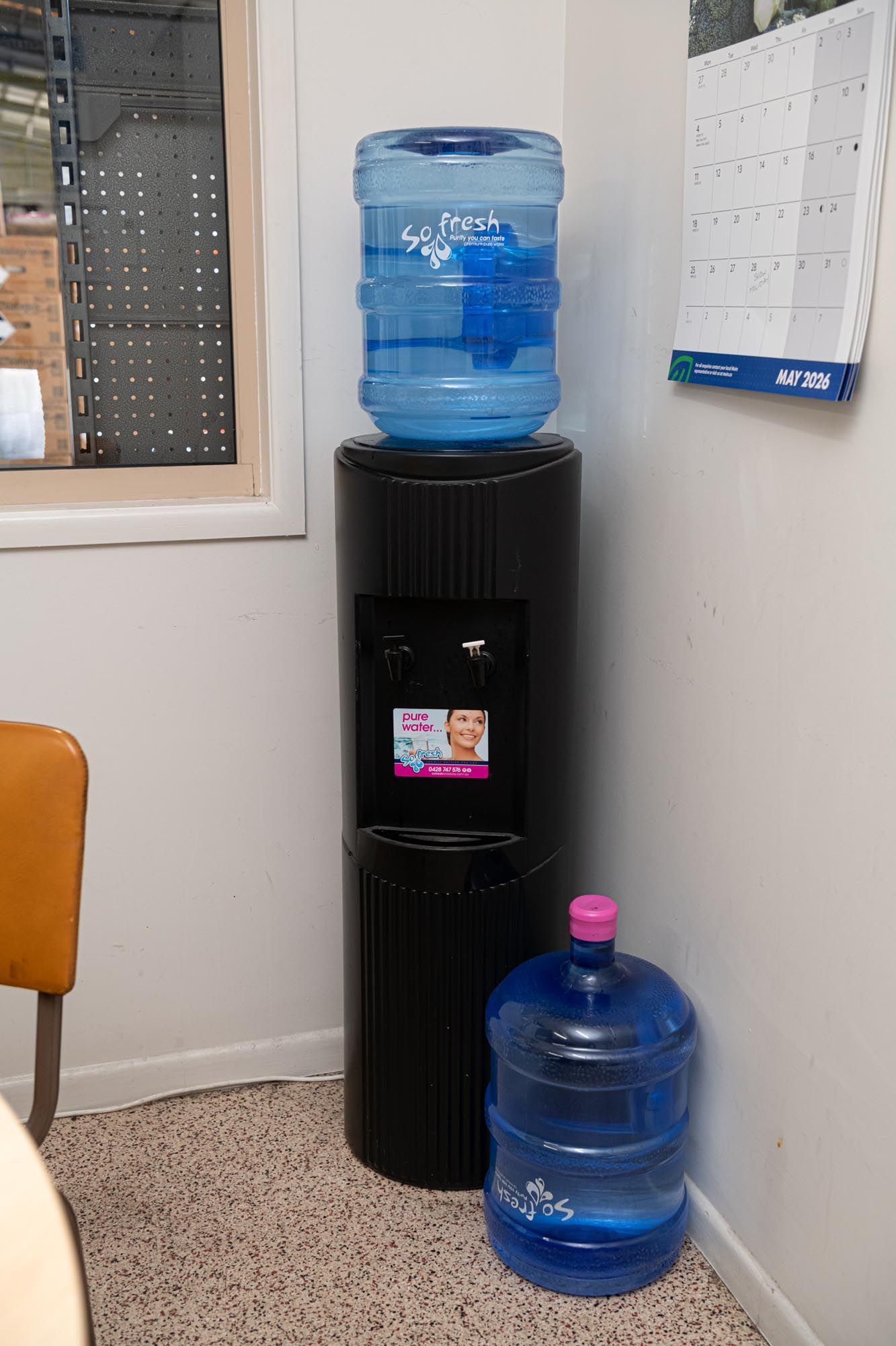 Water cooler with blue jugs in an office corner, next to a wall calendar and window.