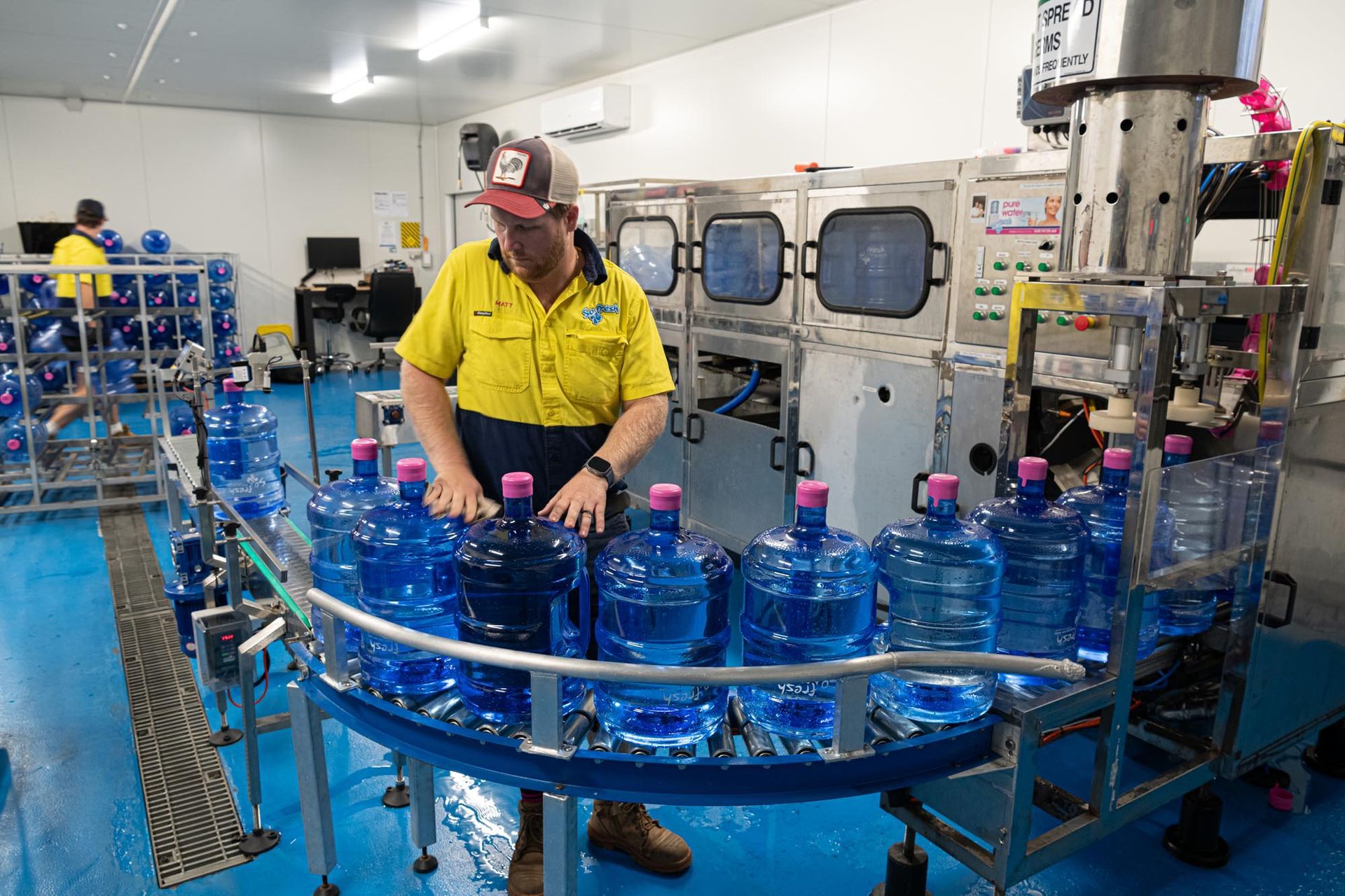 Worker in yellow shirt operating bottling machine beside rows of blue water bottles in a factory