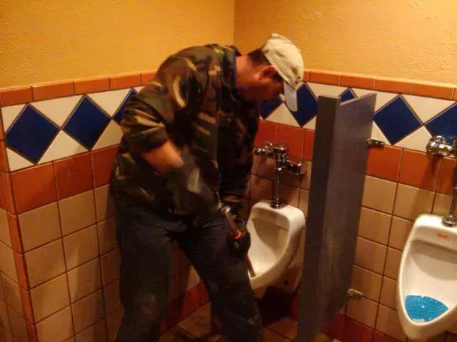 Man in camo shirt working on a urinal in a tiled restroom, with a light-colored hat.