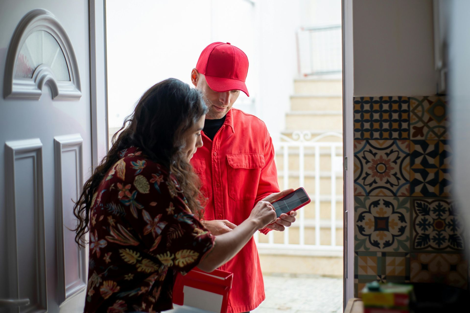 A courier in a red shirt and cap holds a phone for a person to sign for a delivery at a doorway.