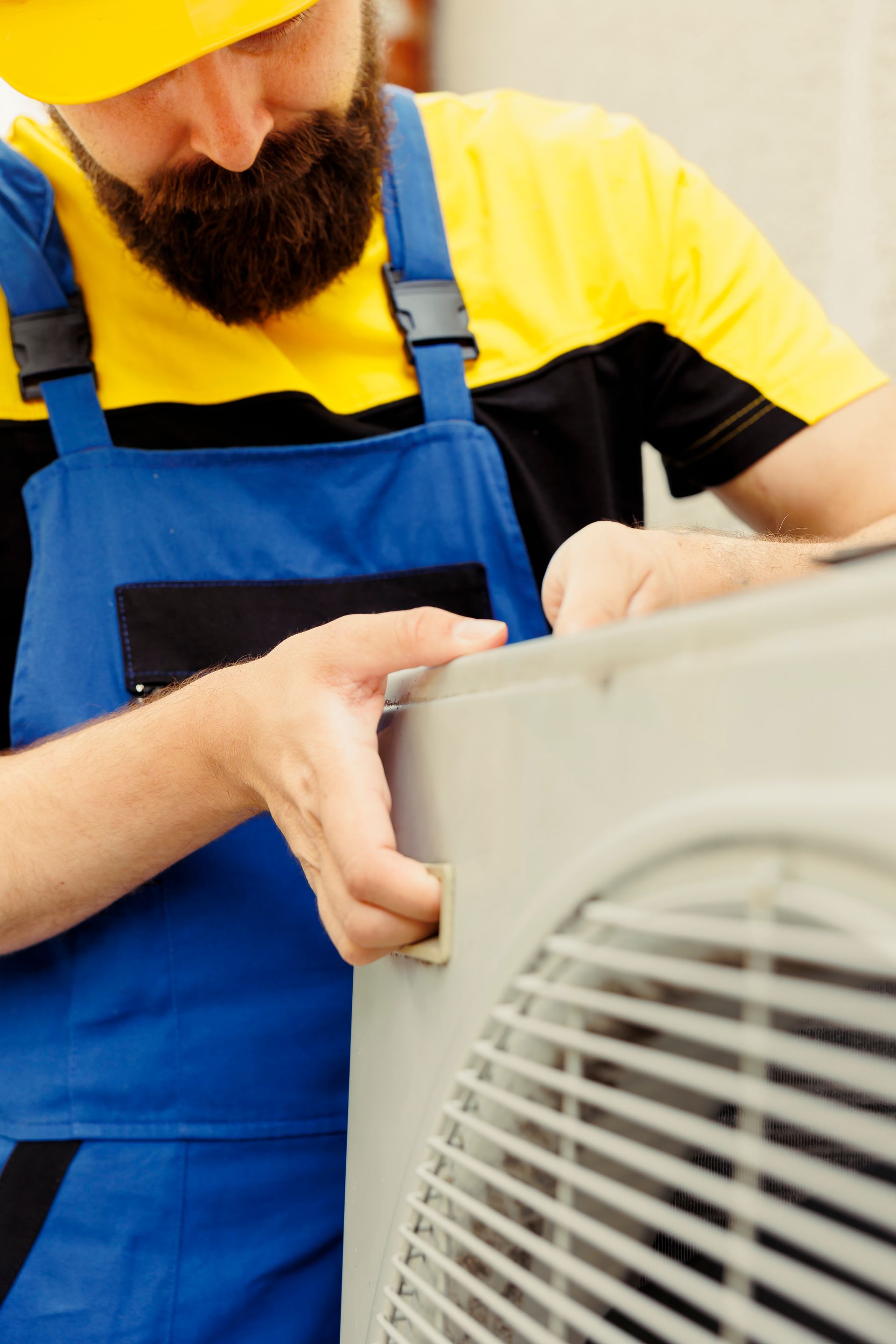 A person in a yellow hard hat and blue overalls works on the metal panel of an outdoor air conditioning unit.
