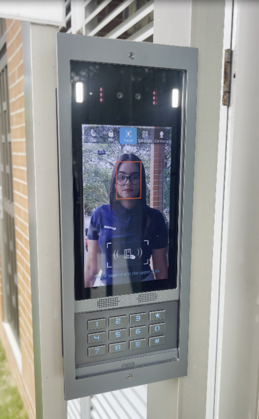 Una pareja señala un timbre con cámara que muestra a una persona sonriente. Están de pie junto a la puerta principal.