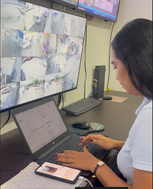 Hombre con camisa blanca y cuello negro mirando la pantalla de un ordenador, inclinado sobre un escritorio con un cuaderno.
