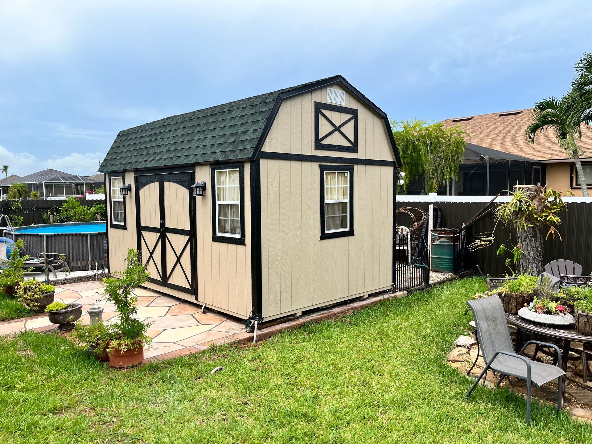 Tan and black barn-style shed in a backyard. Green roof, small windows, and a grassy lawn surround it.